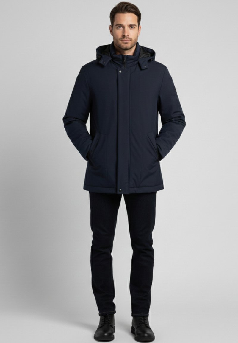 Parka aislante azul marino con capucha, cierre de cremallera, dos bolsillos laterales y puños elásticos, usada sobre pantalones oscuros y botas negras.