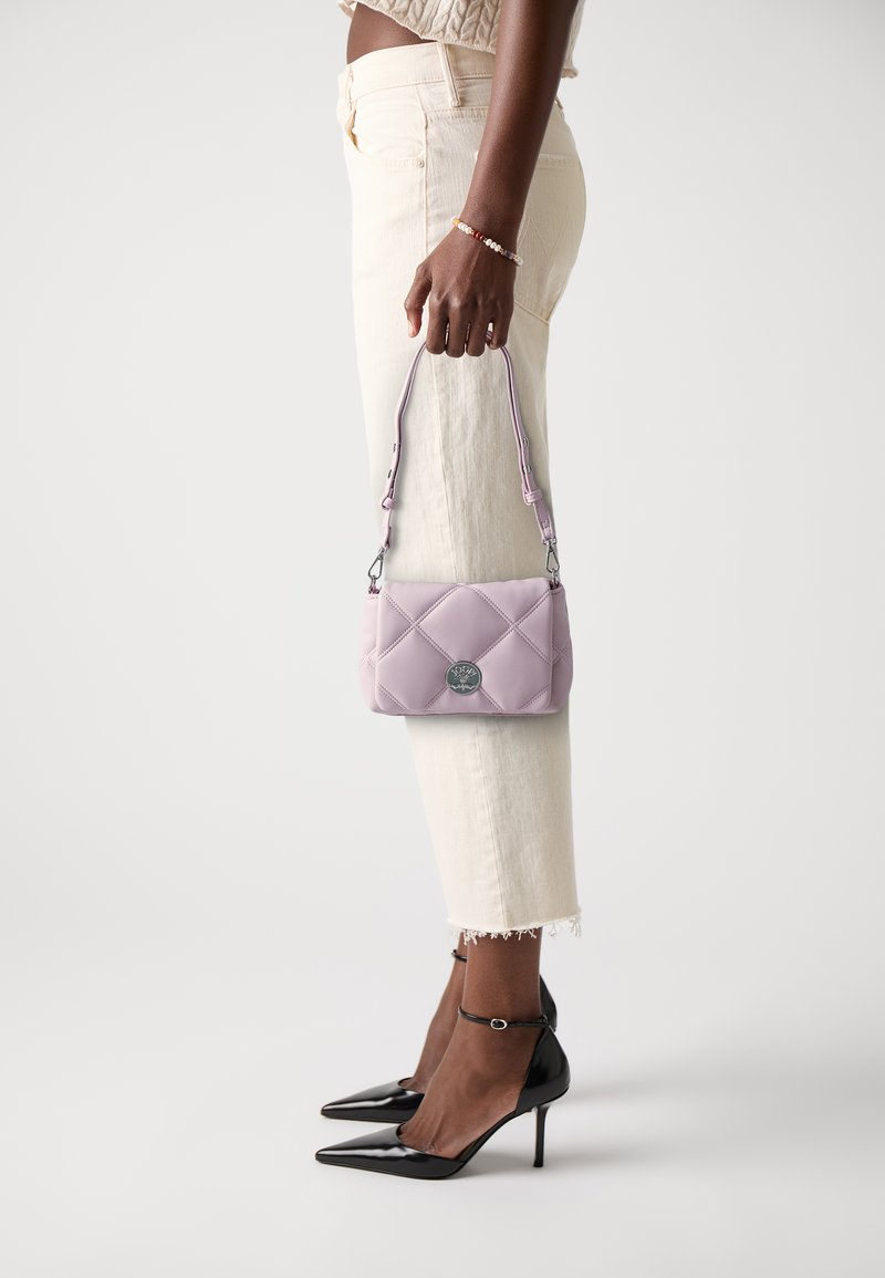 Quiltad lila handväska med en silverlogotyp, hålls av en person som bär krämfärgade jeans och svarta spetsiga klackar. Handväskan har en axelrem.