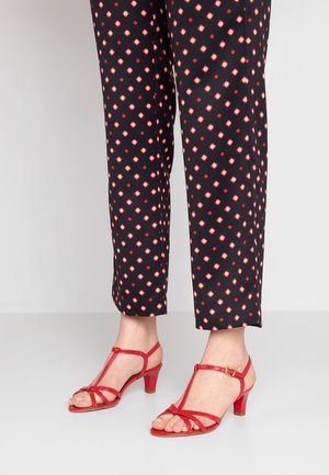 Feet wearing red strappy low-heeled sandals paired with black cropped pants featuring a red and white diamond pattern.