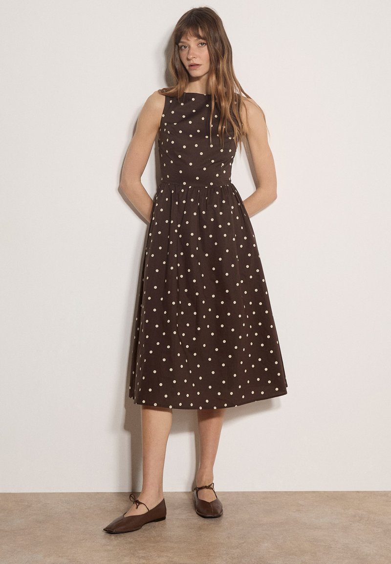 Femme en robe midi marron sans manches avec des pois blancs, debout contre un mur blanc, portant des chaussures plates marron.