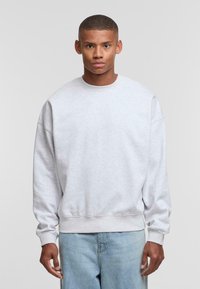 Grauer Sweatshirt mit überschnittenen Schultern, gerippten Bündchen und Saum, weiche Textur, getragen über hellblauen Jeans, einfarbiger Hintergrund.