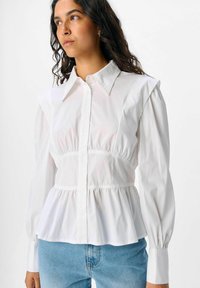 Witte blouse met lange mouwen, een getailleerde taille, gerimpelde schouders en knoopsluiting. Glad stof met een gestructureerd ontwerp en een subtiele glans.