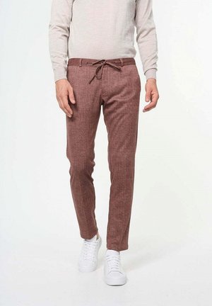 Homme portant un pantalon marron ajusté avec une taille à cordon, un haut beige à manches longues et des baskets blanches, debout sur un fond blanc.