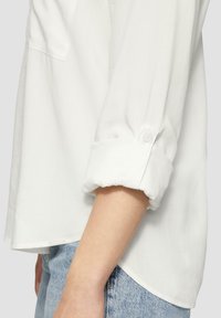 Chemise blanche en tissu léger, avec des manches retroussées et des boutons, et un ourlet arrondi. Coupe décontractée, associée à un jean bleu clair.