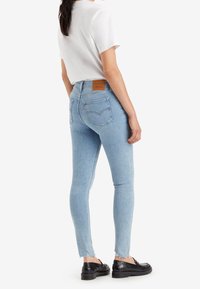 Helle blaue Skinny-Jeans mit fransigem Saum und Gesäßtaschen, kombiniert mit einem weißen kurzärmeligen Oberteil und schwarzen Schuhen. Ein beigefarbener Lederlabel ist sichtbar.