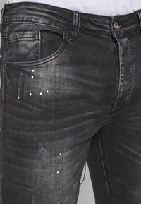 Černé džínové kalhoty z denimu s úzkým střihem, mírným leskem a oprýskanými detaily s bílými kapkami barvy na přední straně. Viditelné kontrastní švy.