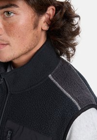 Black fleece vest with a high collar, featuring a textured fabric, a gray shoulder panel, and a zippered pocket. Contrast stitching detail visible.