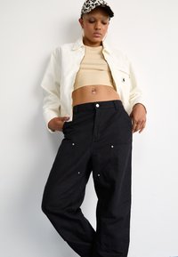 Chaqueta ligera blanca, top corto beige y pantalones negros holgados con bolsillos. La gorra tiene un diseño de estampado de leopardo.