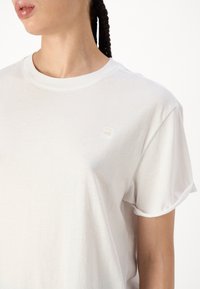 Biała bawełniana koszulka z okrągłym dekoltem, krótkimi rękawami oraz małym wypukłym logo na górnej lewej części klatki piersiowej. Miękka tekstura.