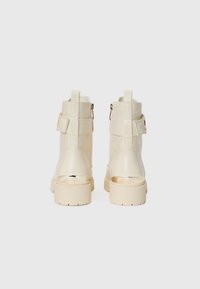 Botas tobilleras color crema con un acabado texturizado, que cuentan con un cierre de cremallera, detalles de hebilla y una suela gruesa de color claro.