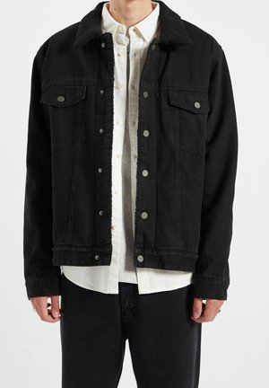 Homme portant une veste noire boutonnée avec un col en sherpa sur une chemise blanche boutonnée et un pantalon noir, debout devant un fond blanc.