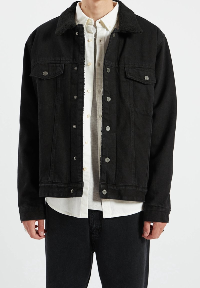 Homme portant une veste noire boutonnée avec un col en sherpa sur une chemise blanche boutonnée et un pantalon noir, debout devant un fond blanc.