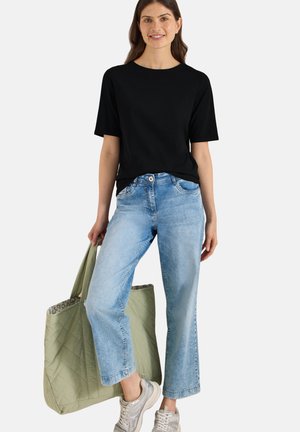 Kobieta uśmiecha się, nosi czarną koszulkę z krótkim rękawem, jasnoniebieskie dżinsy, srebrne trampki i trzyma dużą oliwkowozieloną pikowaną torbę.