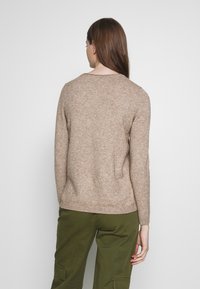 Kvinna med långt brunt hår som bär en beige stickad tröja och olivgröna byxor, stående mot en enkel vit bakgrund.