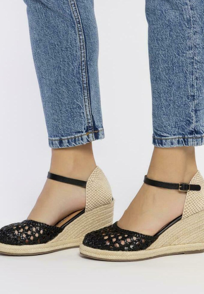 Sandales compensées en tissu noir tressé avec une semelle en paille beige et une bride de cheville noire. Elles présentent un bout ouvert et des panneaux latéraux texturés.