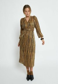 Gouden en zwart gestreepte lange jurk met een aansluitende taille en pofmouwen, gemaakt van een gestructureerde stof. Gestyled met puntige zwarte schoenen.