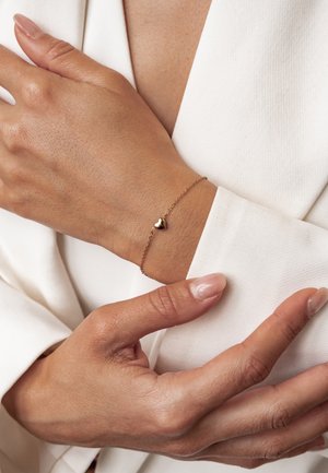 Woman's hand wearing a delicate gold bracelet with a small heart charm over a white long-sleeve garment.