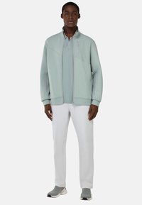 Giacca zip-up verde chiaro con colletto alto, abbinata a un polo coordinato e pantaloni bianchi a sigaretta, completata da scarpe grigie slip-on.