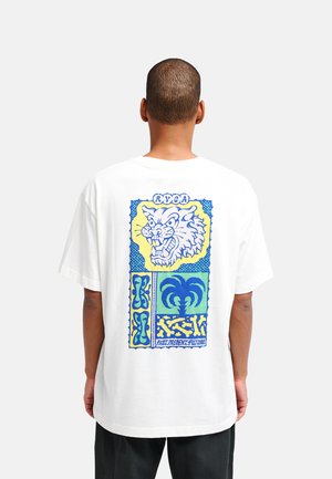 Persona que lleva una camiseta blanca con un gráfico colorido de un gato gruñendo, una palmera y formas abstractas en azul y amarillo en la espalda.