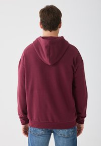Un sweat à capuche bordeaux avec un design au dos, doté d'une grande capuche, de poignets côtelés et d'une taille côtelée, associé à un jeans bleu clair.