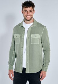 Green textured shirt with buttoned pockets, open collar, and front buttons, layered over a white t-shirt, paired with dark jeans.