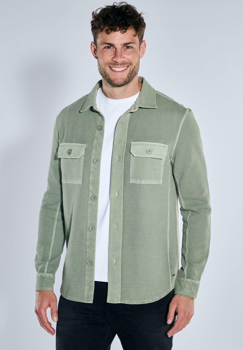 Green textured shirt with buttoned pockets, open collar, and front buttons, layered over a white t-shirt, paired with dark jeans.