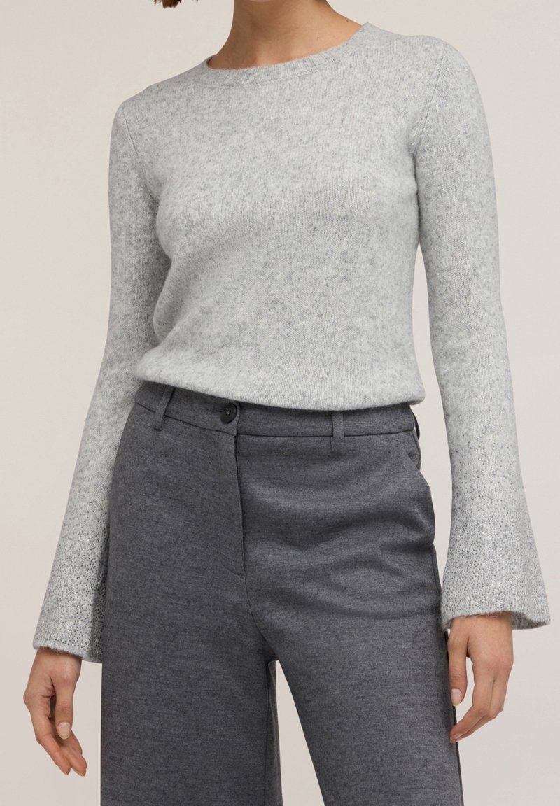 Femme portant un pull en tricot gris clair à manches longues avec des poignets évasés et un pantalon sur mesure gris foncé taille haute.