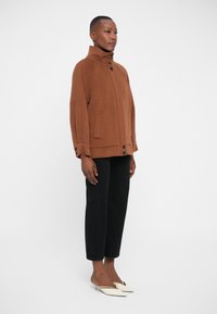 Giacca marrone in misto lana con colletto alto, tasche laterali e chiusura a bottoni; abbinata a pantaloni neri a tre quarti e scarpe slip-on color crema.