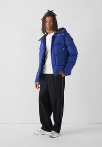 Blauwe pufferjack met een capuchon, voorzien van ritssluitingen en zwarte accenten. Gepaard met een wit shirt en zwarte wijde broek, aangevuld met grijze sneakers.