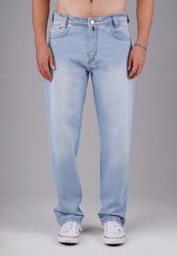 Mann in hellblauen Jeans und weißen Sneakers, mit entspannten Händen an den Seiten vor einem schlichten grauen Hintergrund stehend.