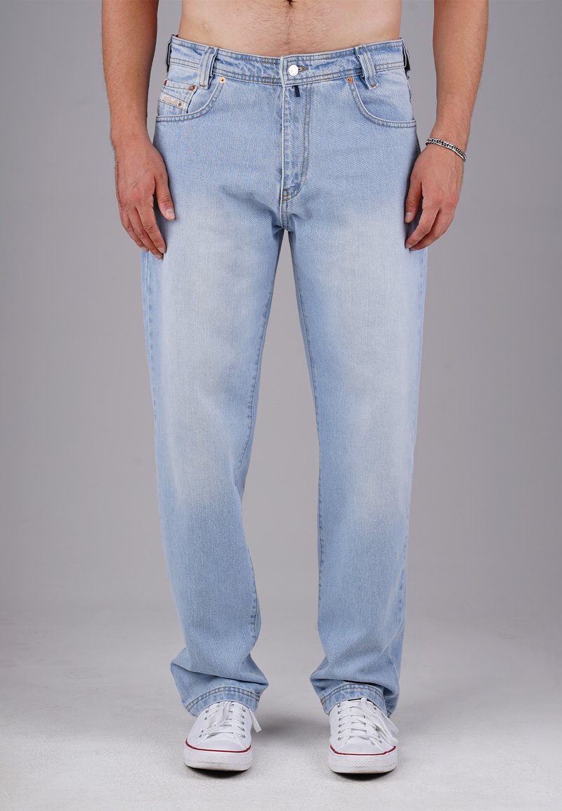 Mann in hellblauen Jeans und weißen Sneakers, mit entspannten Händen an den Seiten vor einem schlichten grauen Hintergrund stehend.