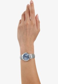 Silbernes Metall-Armbanduhr mit einem runden blauen Zifferblatt, silbernen Stundenmarkierungen und einem Edelstahlarmband. Die Hand ist erhoben und zeigt die Uhr.