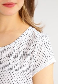 Hvid t-shirt med marineblå geometriske mønstre; korte ærmer, bred halsudskæring og en blød tekstur. Enkel, moderne design med fokus på print.