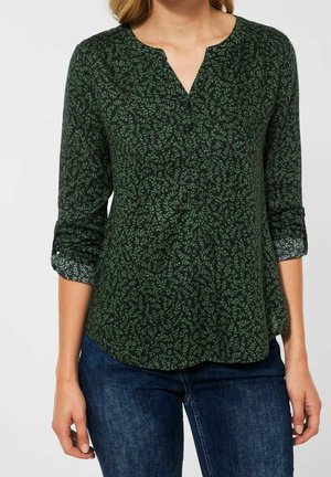 Kobieta ubrana w ciemnozieloną bluzkę z małym wzorem liści oraz rękawami podwiniętymi do połowy przedramienia, w połączeniu z niebieskimi dżinsami.