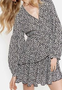 Svart klänning med vitt blommönster, v-ringning, långa puffärmar, lager på lager med volangkant, mjukt tyg och subtil elastikdetalj vid ärmslut.