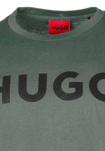 Зелена памучна тениска с кръгло деколте, смела черна надпис "HUGO" на гърдите и червена етикетка на марката на яката.