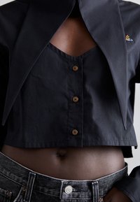 Black cropped jacket with a large collar, featuring brown buttons. Layered over a dark fabric top, with textured dark jeans visible.