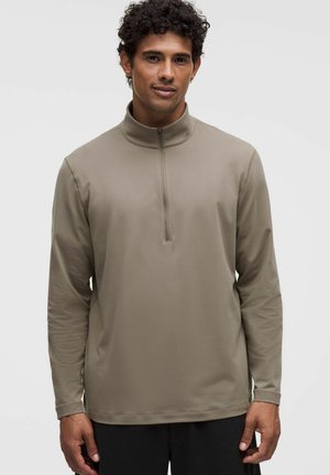 Uomo che indossa un maglione beige a maniche lunghe con una corta cerniera frontale e un colletto alto, in piedi davanti a uno sfondo uniforme.