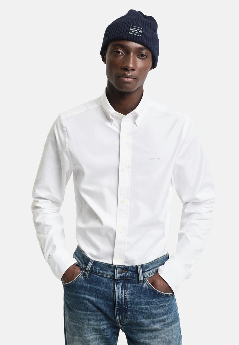 Jeune homme portant un bonnet en tricot marine, une chemise blanche à boutons et un jean bleu, debout les mains dans les poches devant un fond uni.