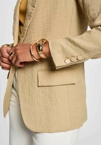 Blazer in lino beige con vestibilità rilassata, tasca a toppa e dettagli con bottoni. Completato da braccialetti dorati sul polso.