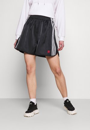 Osoba nosząca czarne szorty Adidas z białymi paskami i czerwonym logo, białą bluzę z kapturem, białe skarpetki oraz czarno-białe sneakersy.