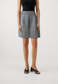 Jupe A-line en tweed gris et blanc avec un motif texturé, longueur mi-cuisse, associée à des sandales à talons carrés noires brillantes.