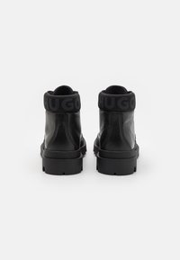 Schwarze Lederstiefeletten mit strukturiertem Finish, runder Spitze, dicker schwarzer Sohle und einem gefütterten schwarzen elastischen Kragen mit dem Aufdruck "HUGO" in fetter Schrift.