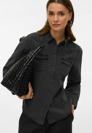 Černá džínová košile s dlouhými rukávy, zapínáním na knoflíky vpředu, dvěma kapsami na prsou a volným střihem. Drží černou tkanou kabelku s texturou.