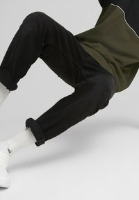 Pantalones negros de corte relajado con puños enrollados, combinados con zapatillas blancas y calcetas blancas de canalé. La parte superior tiene un diseño en oliva y negro.