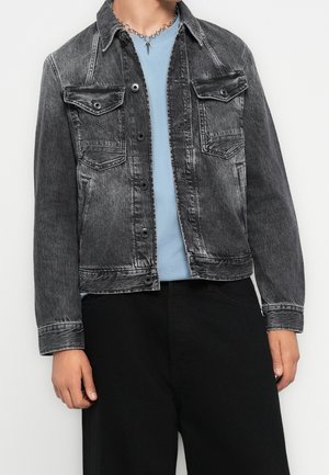 Person trägt eine ausgewaschene schwarze Jeansjacke über einem hellblauen Hemd, dazu schwarze Hosen und eine silberne Kettenkette.