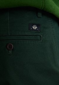 Mørkegrønne bomullsbukser med en glatt tekstur, med knappelukking og et logo-patch på baklommen.