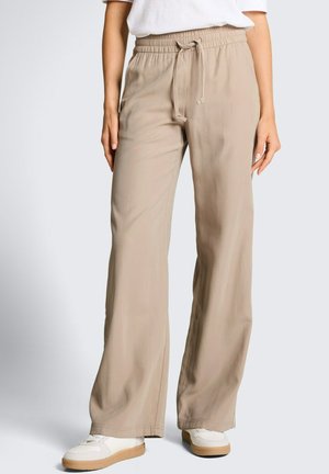 Kobieta w beżowych szerokich spodniach z elastycznym pasem i sznurkiem, białych trampkach i białej bluzce, stoi na tle jednolitego tła.