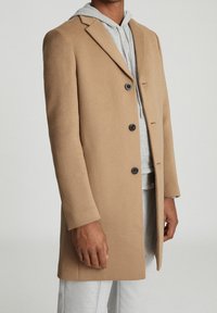 Camel wollen mix jas met een getailleerde pasvorm, met klepjes, vier zwarte knopen en zijzakken, gelaagd over een grijze hoodie.
