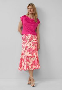 Haut drapé rose et une longue jupe évasée à motif floral dans des nuances de rose et de crème. Des sandales blanches à lanières complètent la tenue.